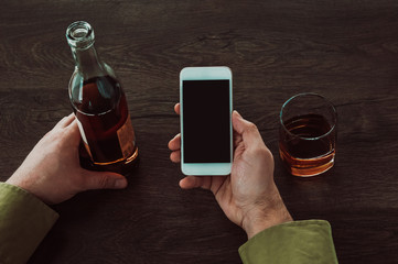 A man holds a mobile phone in his hands. Next on the table is a glass and a bottle of whiskey.