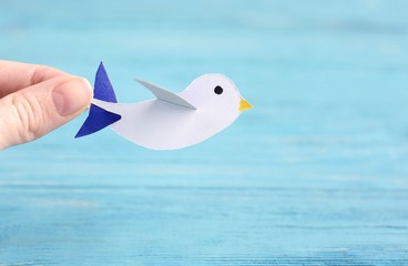 White female hand holding beautiful paper bird with blue wings and yellow beak on blue wooden textured background. Spring symbol creative origami birds. Paper cut animal 