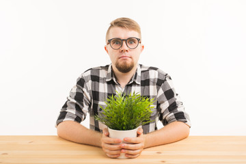 Student, office and people concept - a young grumpy man sitting at the table with a green plant on white background. He looks serious and strange
