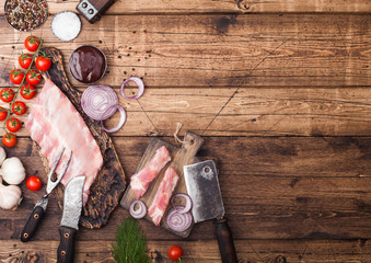Fresh raw pieces of pork ribs on chopping board and vintage meat hatchet, fork and knife on wooden background. Fresh tomatoes and red onion with garlic salt and pepper. Space for text