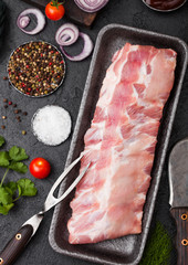 Plastic tray with raw pork ribs and vintage knife and fork on black stone background. Fresh tomatoes and red onion with garlic salt and pepper.