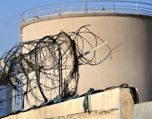 Tank im Industriehafen von Manama mit stacheldrahtgeschützter Mauer im Vordergrund