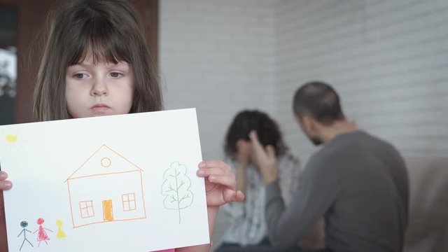 Family problems. Divorce. Sad little girl with a photo of a family against the background of parents quarreling with each other.