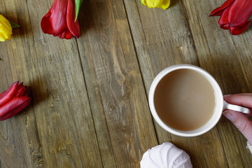 Flat lay of greeting card with flowers tulips and hand keeping white cup of coffee with milk on brown wooden background. Top view. Copy space