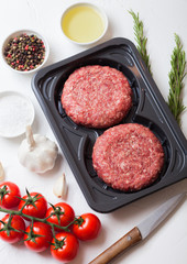 Plastic tray with raw minced homemade beef burgers with spices and herbs. Top view. On top of white kitchen table background with tomatoes salt and pepper.