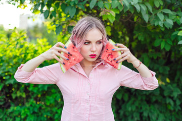 Beautiful young woman with pink hair holding two slices of watermelon in front of her face