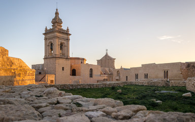 Fototapeta na wymiar Side view on the old, historical St. Joseph's Chapel inside the Citadel of Victoria surrounded by antique ruins, walls with grass field and sandstones in the foreground on Gozo, Malta.
