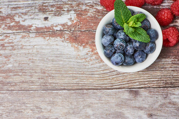 top view on blueberries in a white bowl with some peppermint on a wooden background, some raspberries in the corner, with copy space