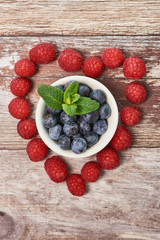 raspberries on wooden surface in a heart shape, there are blueberries in a white bowl in the middle of the heart