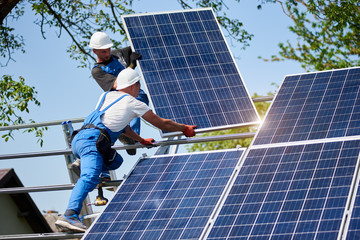 Two workers mounting heavy solar photo voltaic panel on tall steel platform on green tree and blue sky background. Exterior stand-alone solar panel system installation, dangerous job concept.
