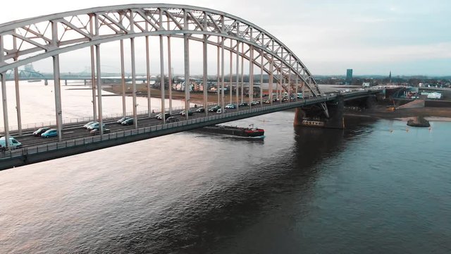 Aerial footage of a transport boat on the river passing under a bridge with traffic. Flying forward and lowering closing in on the boat. This river is 'the Waal' in the Netherlands near Nijmegen.