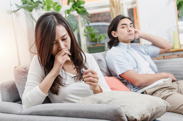 sadness young asian wife crying and looking at her ring sitting on sofa after fight with husband behind her in house interior together, love, divorce couple, family issues and relationship concept