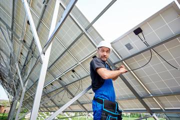 Young engineer technician with funny scared facial expression making electrical wiring standing inside exterior solar panel photo voltaic system on bright sunny summer day. Job and problems concept.