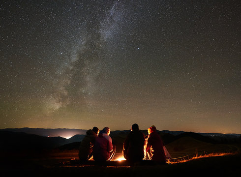 Night summer camping in the mountains. Back view group of four friends tourists sitting on a bench made of logs together around campfire under amazing night starry sky full of stars and Milky way. - Powered by Adobe