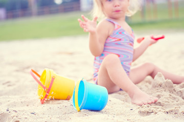 Little blonde baby girl playing with sand on the beach using plastic toys.