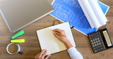 Female hand in a shirt writes in a leather notebook. Computer, cup of coffee, graphics, drawings, calculator on the table in the office. Business and Finance.