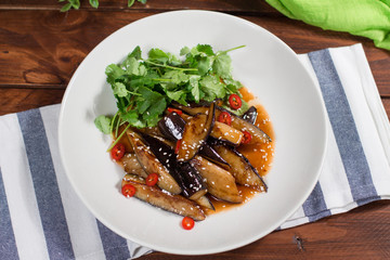 Eggplant slices in spicy red sauce in clay bowl on wooden background. The view from the top.