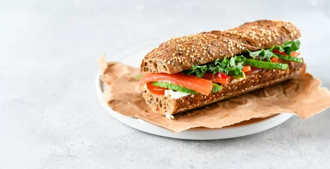 Poster sandwich van een ontbijtgranen stokbrood met avocado, zalm, roomkaas, tomaten en sla bladeren op een witte plaat. lichte achtergrond, selectieve focus en kopieerruimte © KEA