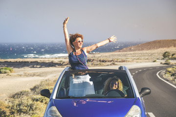 Summer vacation and travel concept image of a young woman out of hood of convertible car. Curly brunette girl arms up, having fun in a cabriolet topless. Female enjoying freedom