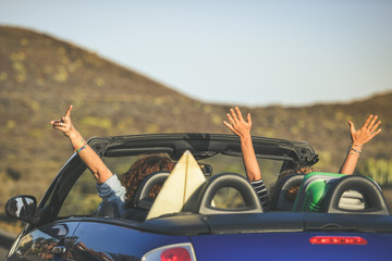 Seen from behind a pair of euphoric women in a convertible car, twisting, waving two curly girls on...