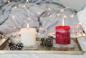 Golden metal tray with two candles of white and red color and decorative pine cones, lights as background. Bedroom decor. Hygge style