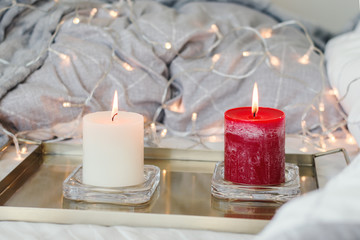 Obraz na płótnie Canvas Two candles of white and red color on golden metal tray, gray blanket and home decor lights as background