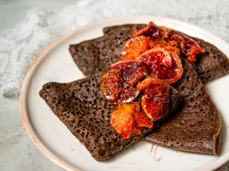 Thin chocolate pancakes with blood oranges sauce on a white plate on gray background with lace fabric. Stack of crepes, russian blin, Maslenitsa. Tasty breakfast. Close up, selective focus