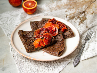 Thin chocolate pancakes with blood oranges sauce on a white plate on gray background with lace fabric. Stack of crepes, russian blin, Maslenitsa. Tasty breakfast. Close up, selective focus