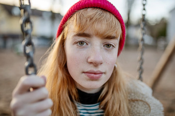 rothaariges Mädchen mit roter Mütze sitzt auf einem Spielplatz auf einer Schaukel