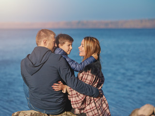 Beautiful European family is sitting on the big stone near the lake, they are happy together and smiling to each other.