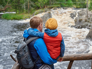  European man and his son are standing on the wooden bridge and enjoying the picturesque view of the wild rough river.