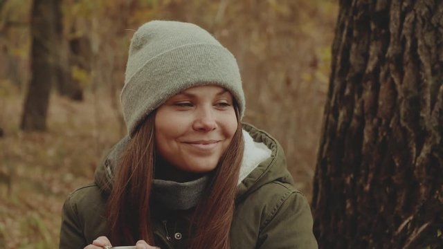 Beautiful happy young woman drinks hot tea near campfire in autumn forest,close-up,oak leaves,near river.Female with her mixbreed dog.slow motion.