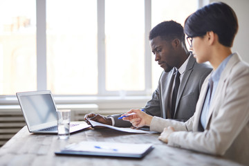 Two young intercultural business employees in formalwear sitting by desk in office and discussing papers