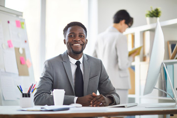 Fototapeta premium Young cheerful African-american businessman in formalwear sitting by table in front of camera during work in office