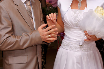 The groom in a wedding suit puts a wedding ring on the bride. She holds a wedding bouquet in another hand.