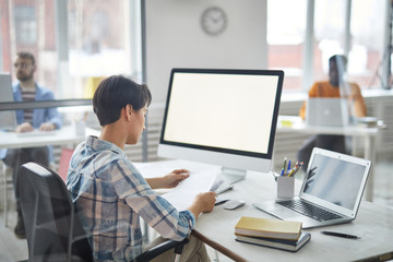Young manager in casualwear looking through business papers while sitting by desk in front of computer and laptop