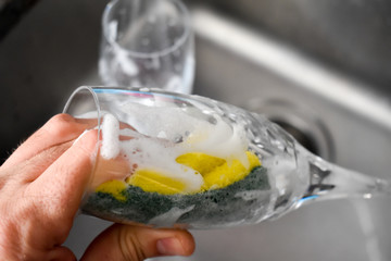 Person washing the glass by hands with a green yellow sponge full of foam in the grey sink at the kitchen and a glass cup just washed