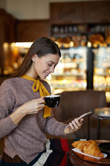 Happy long-haired girl in smart casual having tea or coffee with croissant in cafe and making selfie