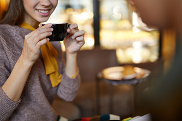 Young smiling elegant woman with cup of tea or coffee sitting in front of camera in cozy cafe
