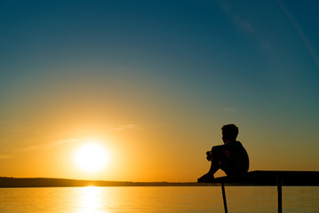 Small boy sitting alone on the footbridge holding his legs and dreaming at sunset. Little kid...