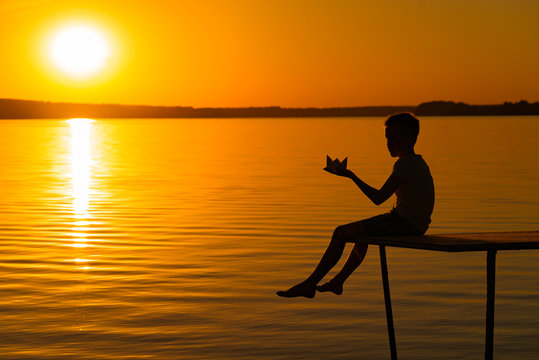 Silhouette Of A Little Boy Sitting On The Bridge And Holding Origami Boat With Bare Legs Over The Water At Sunset. A Child Near The Lake Looks At Beautiful Sunset In Summer.