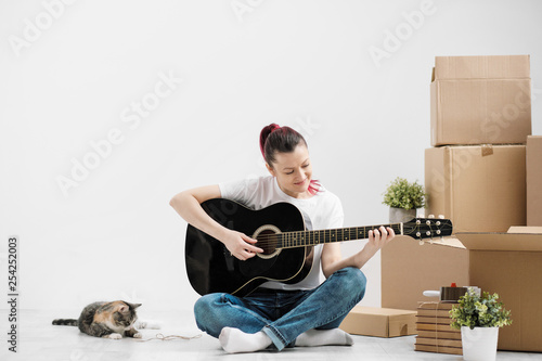 A Young Woman In White T Shirt Is Resting And Playing The Guitar A Young Woman In White T Shirt Is Resting And Playing The Guitar On Background Of Cardboard Boxes New Empty Apartment With Little Pet Cat