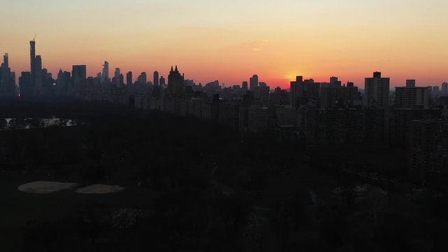 Epic drone slide over the top of New York City's Central Park at gorgeous sunset golden hour.  Rotating view of the Upper East Side skyline while the gorgeous red sun stays as a focal point.  in 4K.