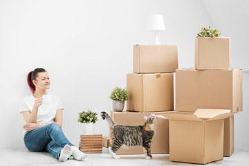 A young beautiful brunette girl in a white T-shirt is sitting on the floor of a bright room and making calls on her smartphone. Around cardboard boxes and a cat.