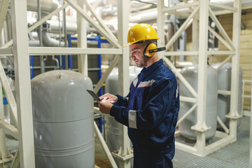 Side view of Caucasian worker in protective suit, helmet and antifones using tablet while standing...