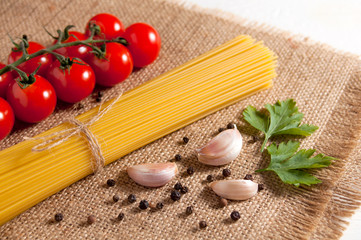 Bunch of raw spaghetti, cherry tomatoes, peppers, garlic cloves and two parsley leaves on sackcloth on a white wooden background. Close-up.
