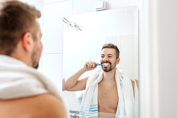 Smiling shirtless man with towel around neck brushing his teeth while standing in front of mirror in bathroom. Morning routine concept.