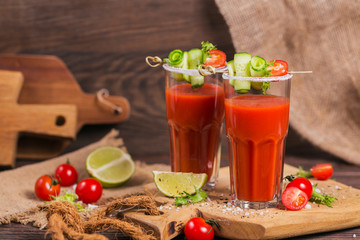 Two glasses of fresh organic tomato juice decorated with raw tomatoes, cucumber and leaves on a rustic wooden cutting board
