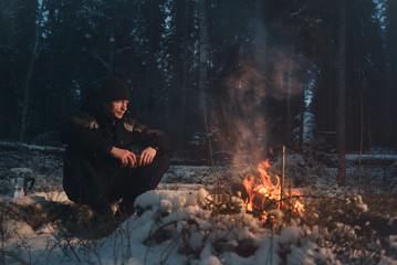 A man sits in the evening winter forest near the fire.
