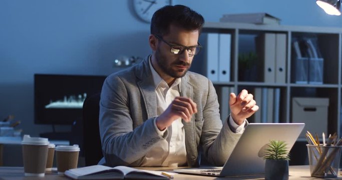 Caucasian young good looking man in glasses working hard late at night at the computer and thinking over difficult task.